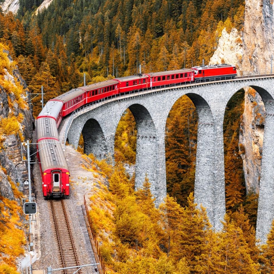 Red train crossing a stone viaduct through forested mountains